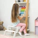 Little Girl Sitting On Step Stool Near Shelf With Toys