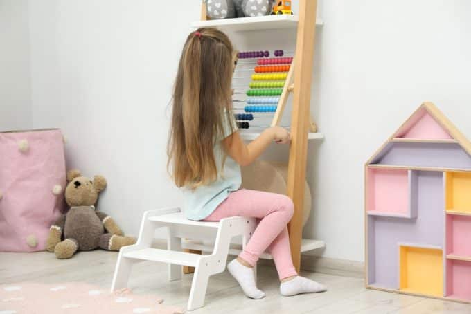 Little Girl Sitting On Step Stool Near Shelf With Toys