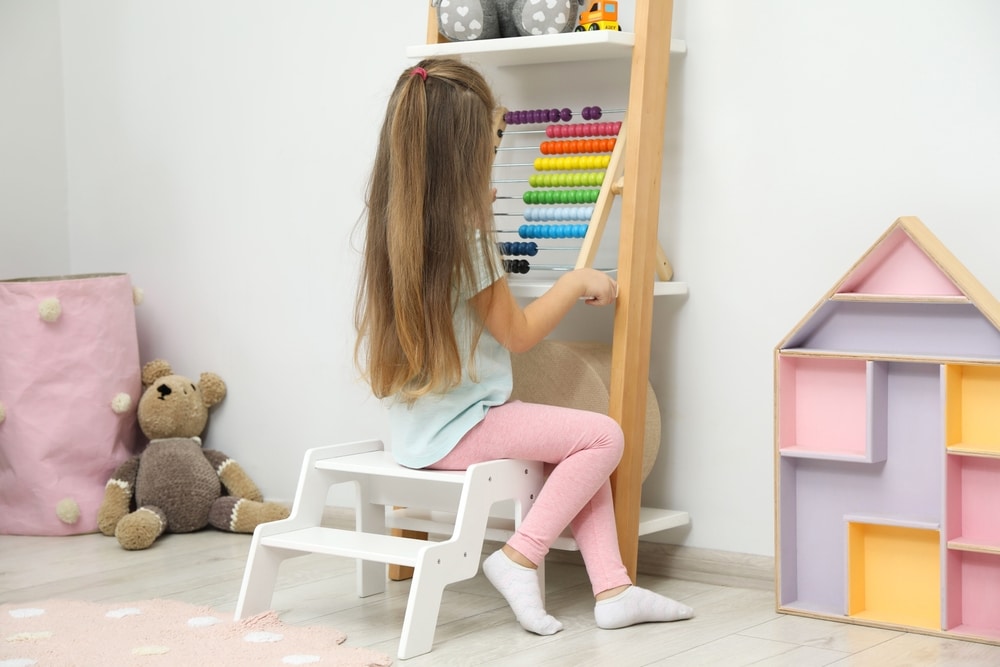 Little Girl Sitting On Step Stool Near Shelf With Toys