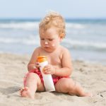a baby sitting on beach holding a bottle of sunscreen