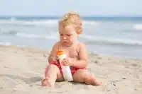 a baby sitting on beach holding a bottle of sunscreen