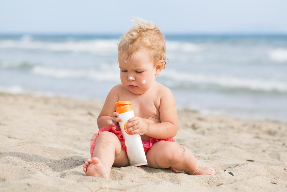 a baby sitting on beach holding a bottle of sunscreen