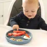 a baby eating food from grey colored divided plate