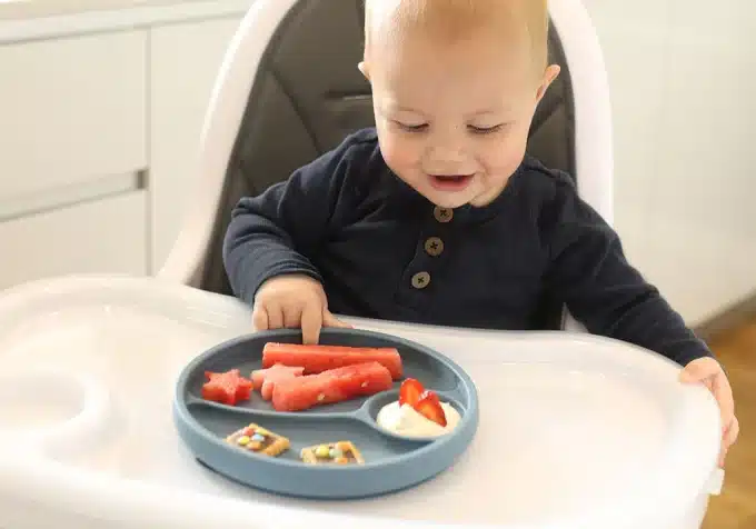 a baby eating food from grey colored divided plate