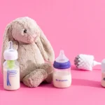 different baby bottles placed on a pink surface with a teddy