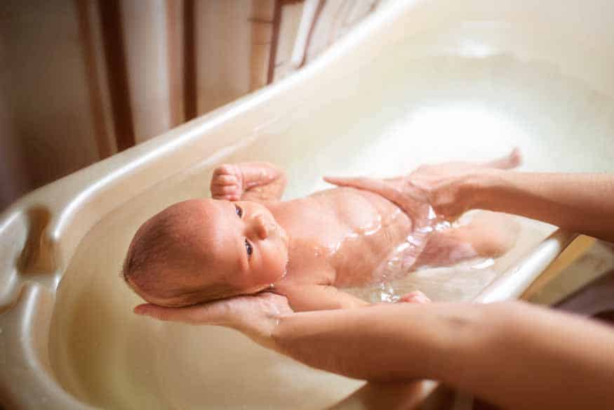 A caregiver gently bathes a newborn baby in a small tub supporting the baby s body with their hands in the warm water
