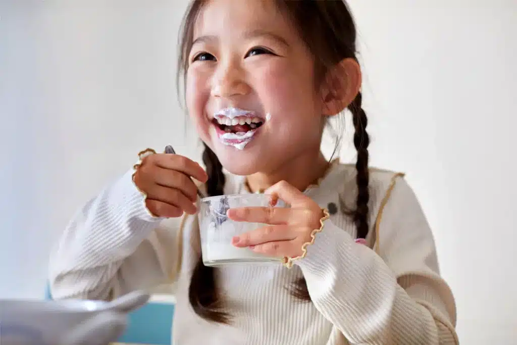 A young girl with braided hair enjoys a bowl of yogurt smiling happily with yogurt on her face