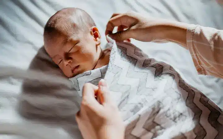 Newborn baby sleeping peacefully while being gently swaddled in a patterned blanket by an adult