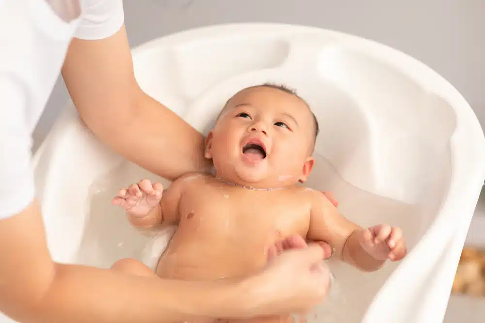 Smiling baby in a small white bathtub being gently bathed by an adult