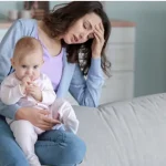 A depressed mom with one hand on her forehead and holding her baby with other