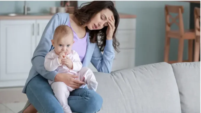 A depressed mom with one hand on her forehead and holding her baby with other
