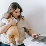 a mom holding her baby working on a laptop