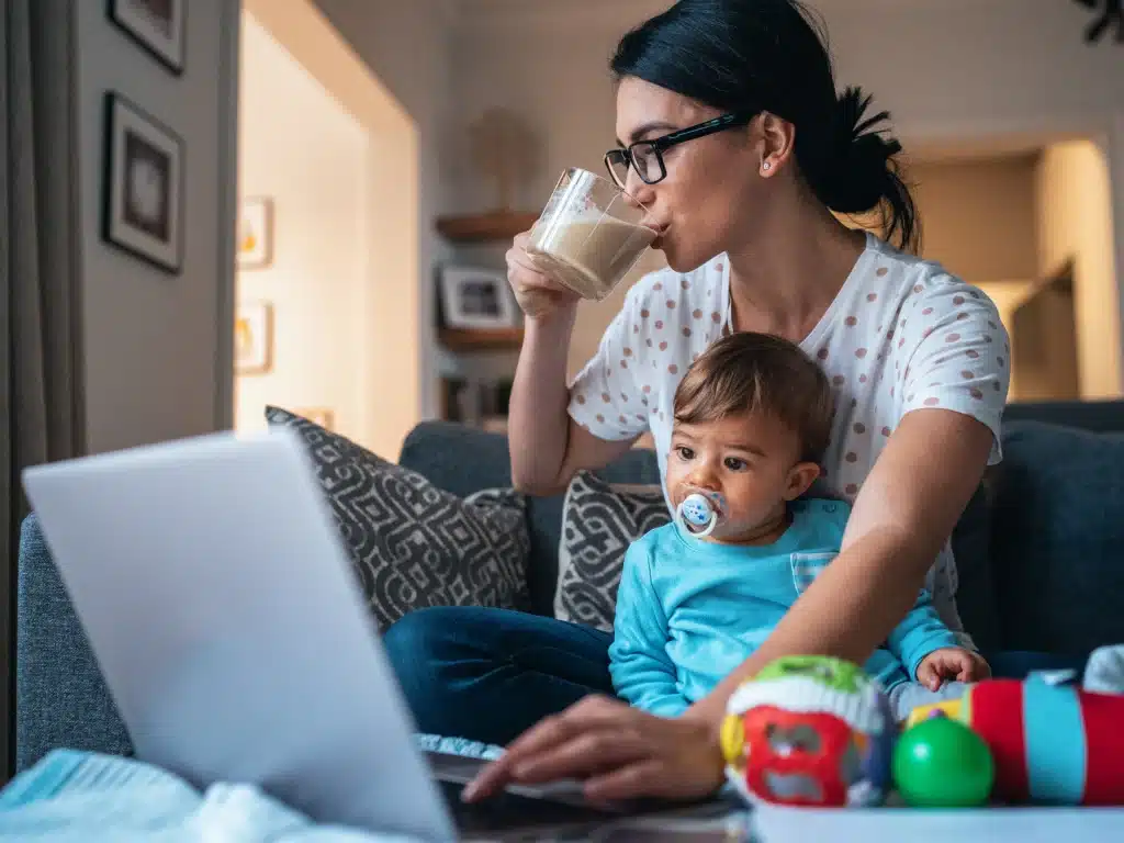 a working mom working on a laptop and sipping coffee a baby in her lap